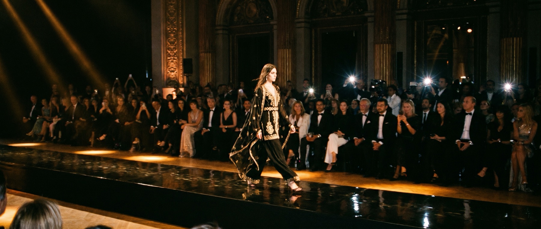 Runway moment at an after-dark fashion show, warm spotlights over a dark room